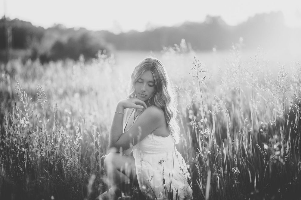 Black and white photo of a young woman in a white dress sitting in a sunlit wildflower field.