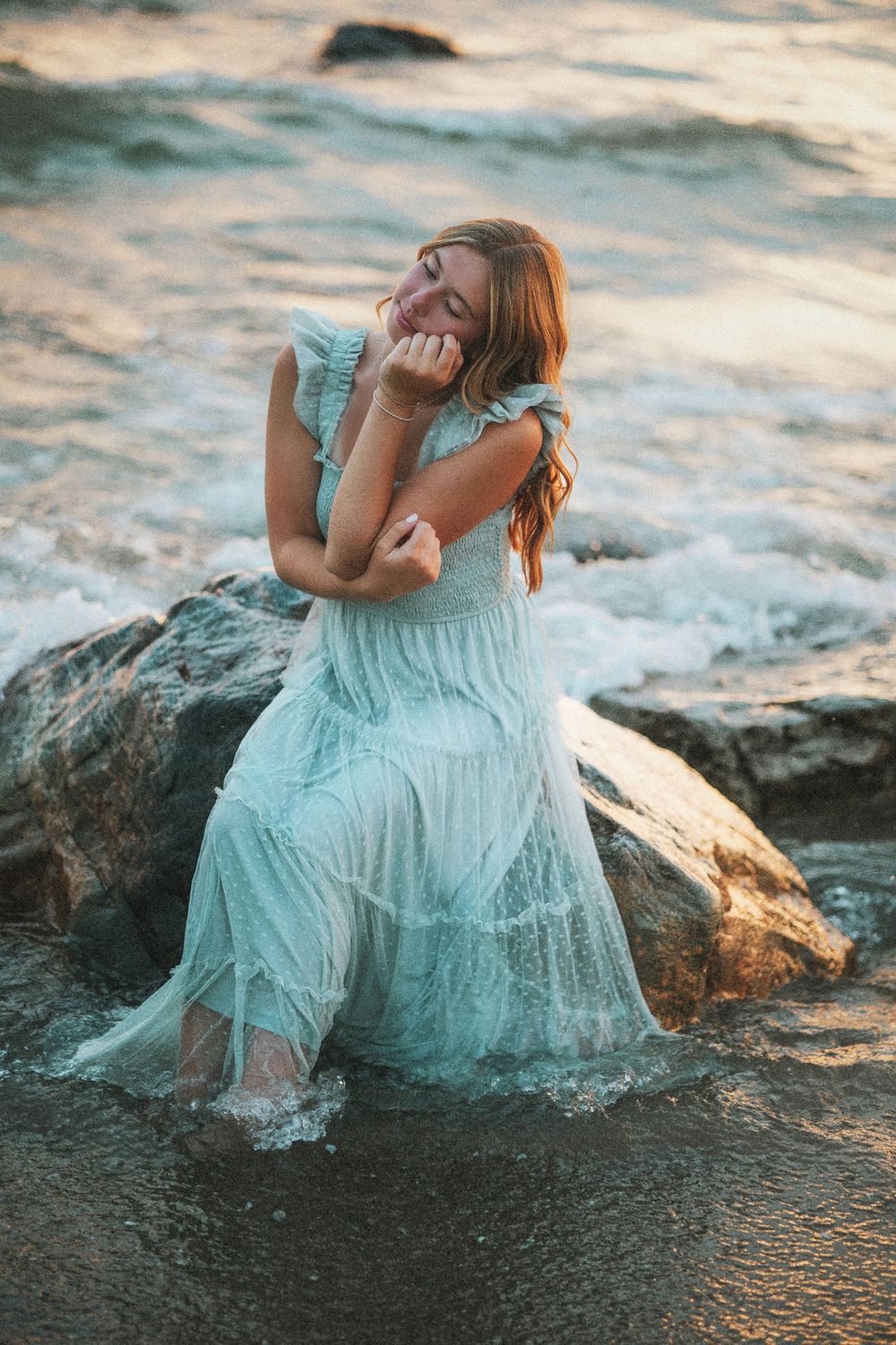 Boyne City senior  in a flowing mint dress sits on rocks by the lake michigan waves at golden hour sunset.