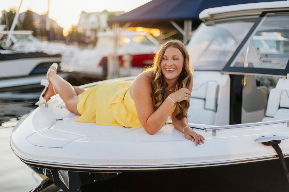 A smiling woman in a yellow dress lying on a white boat at a marina during golden hour sunset.