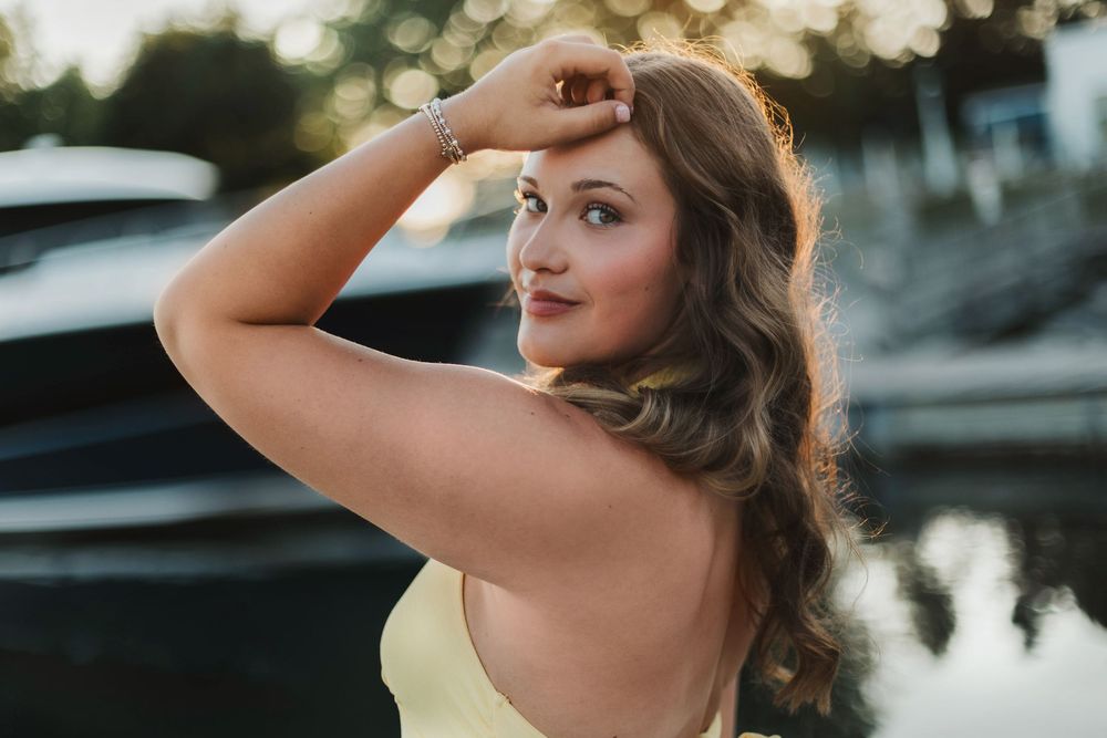 Young woman in yellow dress posing outdoors near water at golden hour with elegant wavy hair.