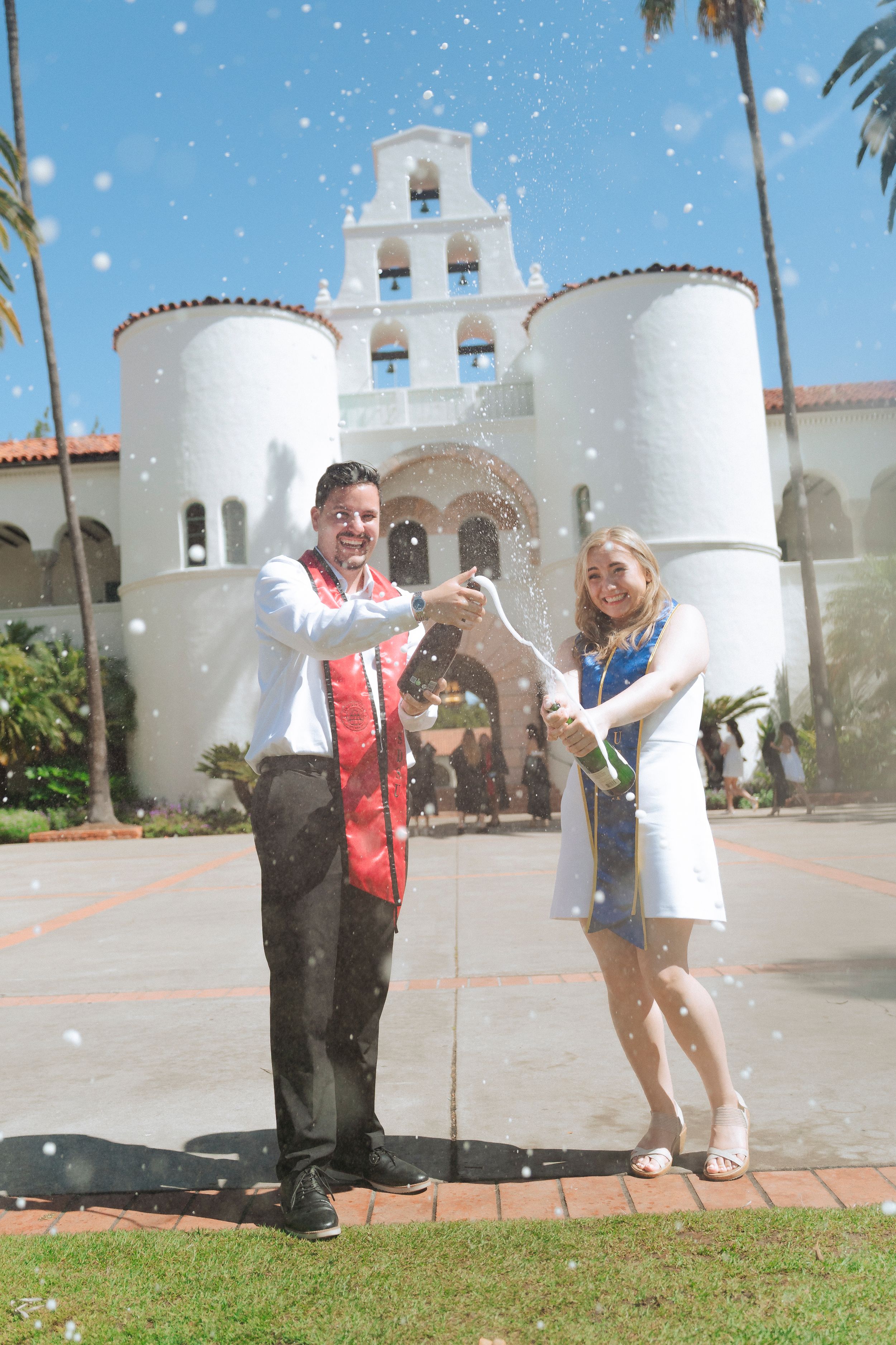Two people pop a bottle of champagne in front of a beautiful backdrop with clear skies.