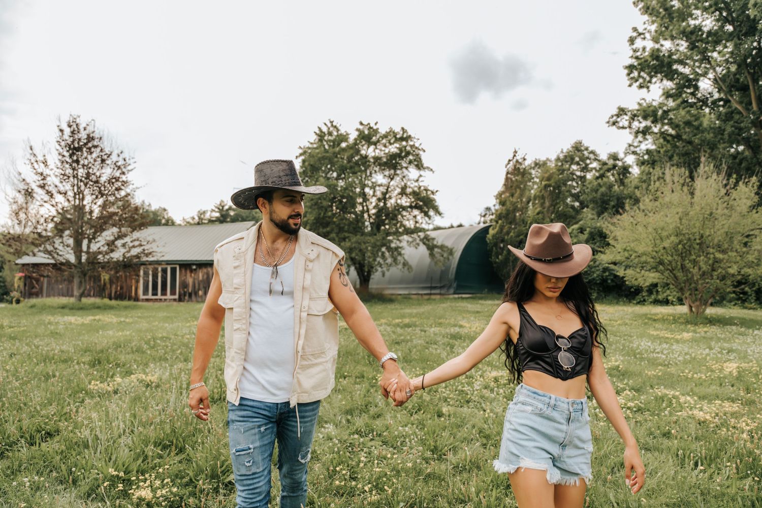 A couple holding hands while walking through a sunny field wearing casual western-inspired outfits.