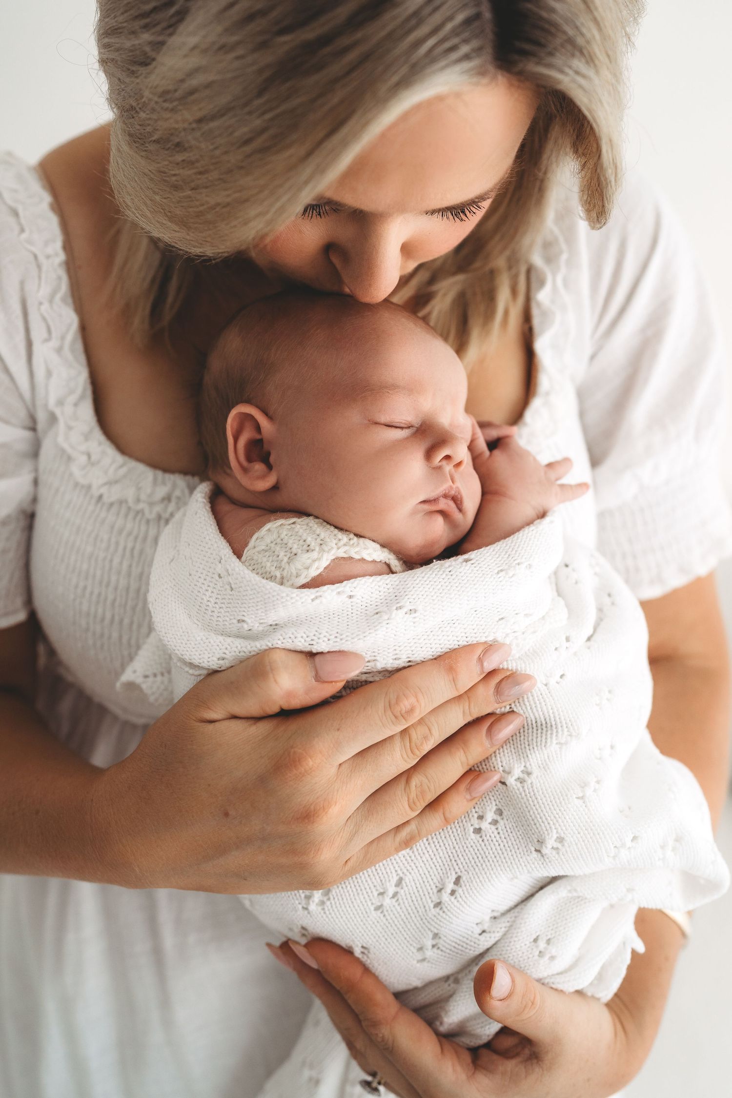 Loving mother with blonde hair kissing sleeping newborn baby wrapped in white knit swaddle blanket.