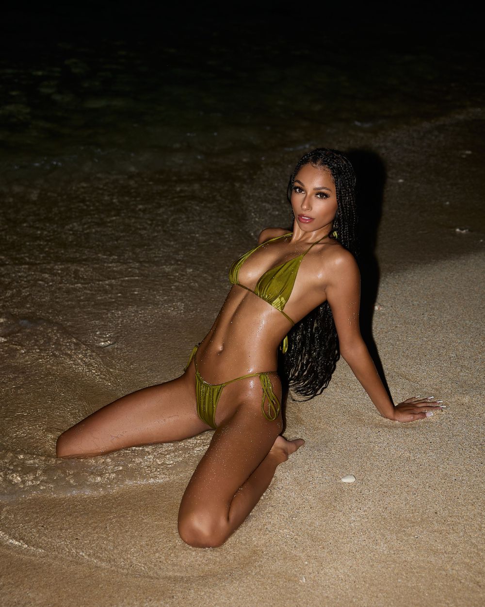 Model in green bikini posing on wet sand at night, with waves gently washing around her under soft beach lighting.