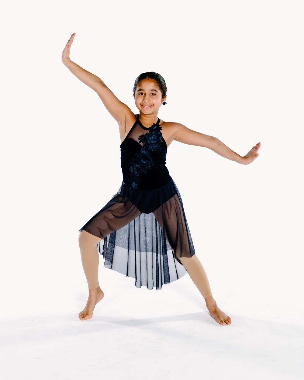 Young dancer in black sequined leotard and sheer tulle skirt performs graceful contemporary dance pose on white background.