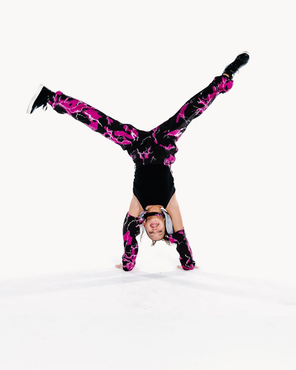 A dancer in a pink and black floral print outfit performs a cartwheel handstand against a white background.
