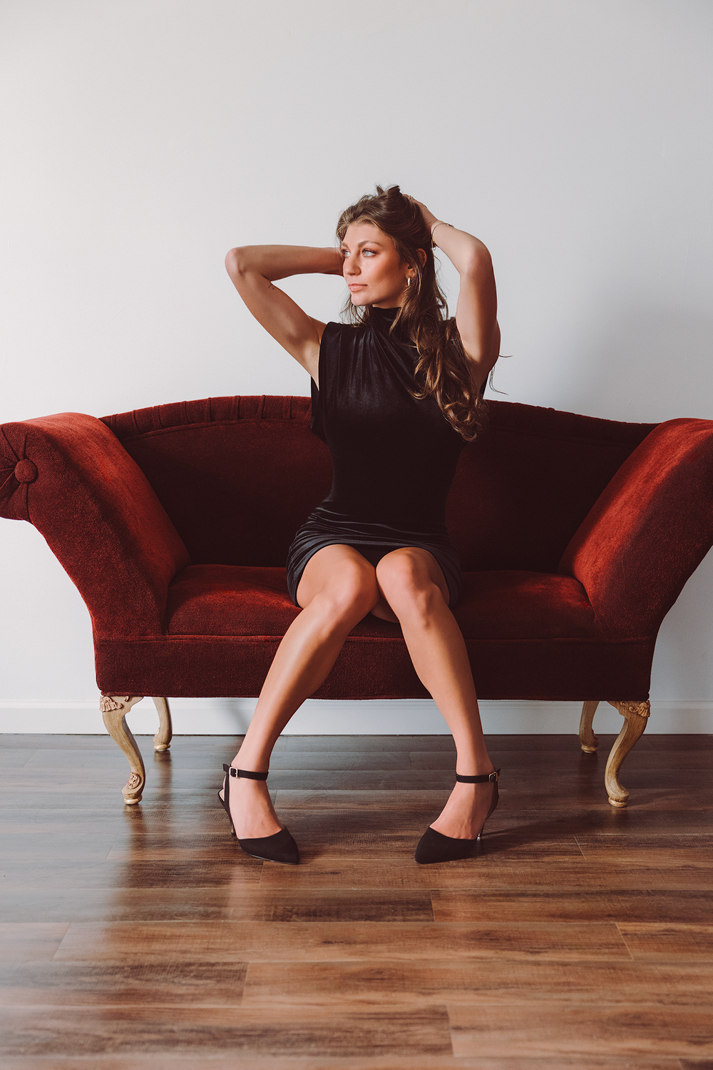 Elegant figure in little black dress poses on vintage red velvet sofa against white wall with hardwood floors.