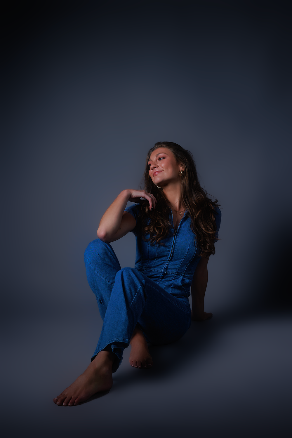 Artistic portrait in moody lighting shows a person in denim outfit sitting on dark floor with dramatic shadows.
