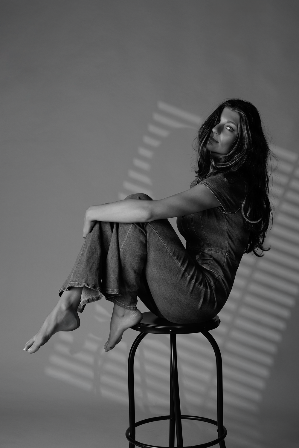 Artistic black and white portrait of a person perched on a stool with dramatic window blind shadows.