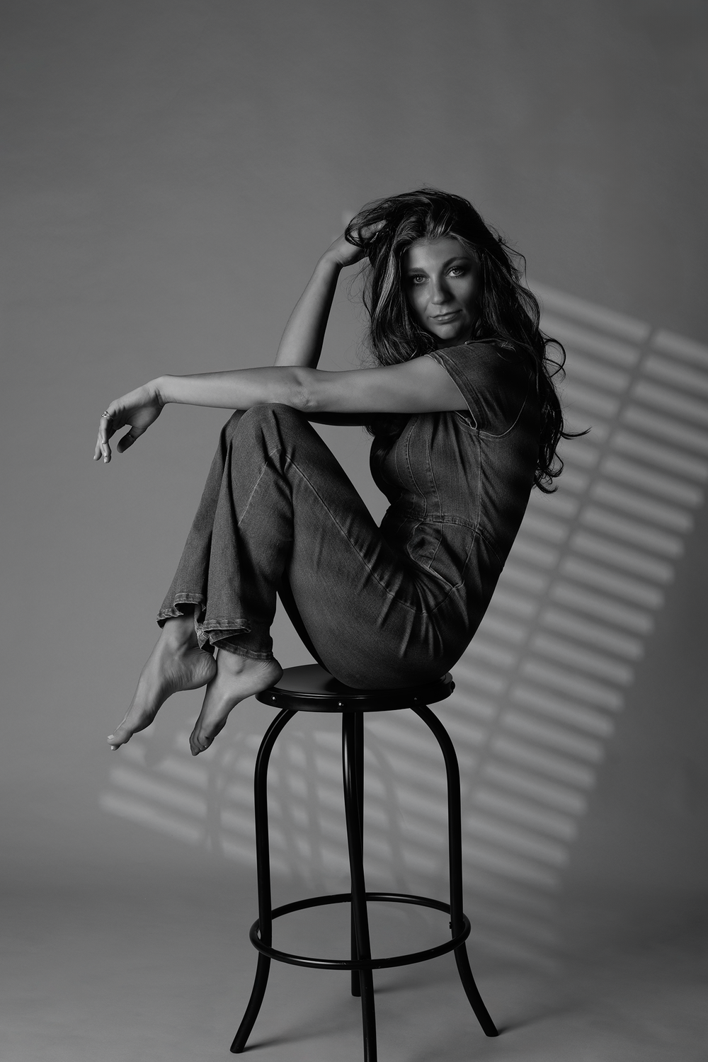 Artistic black and white portrait of a model posing casually on a barstool with window blind shadows in background.