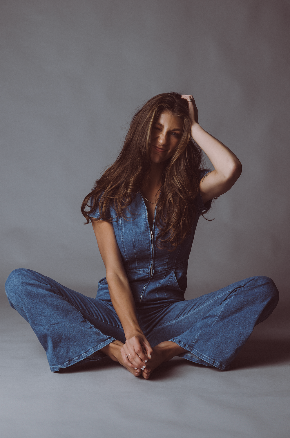 Person in denim outfit sitting cross-legged on floor in casual pose against gray background.