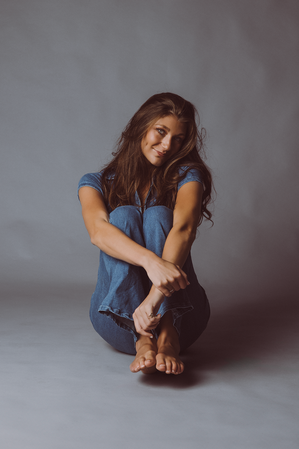 A person in casual denim attire sits cross-legged on a gray floor against a neutral background.