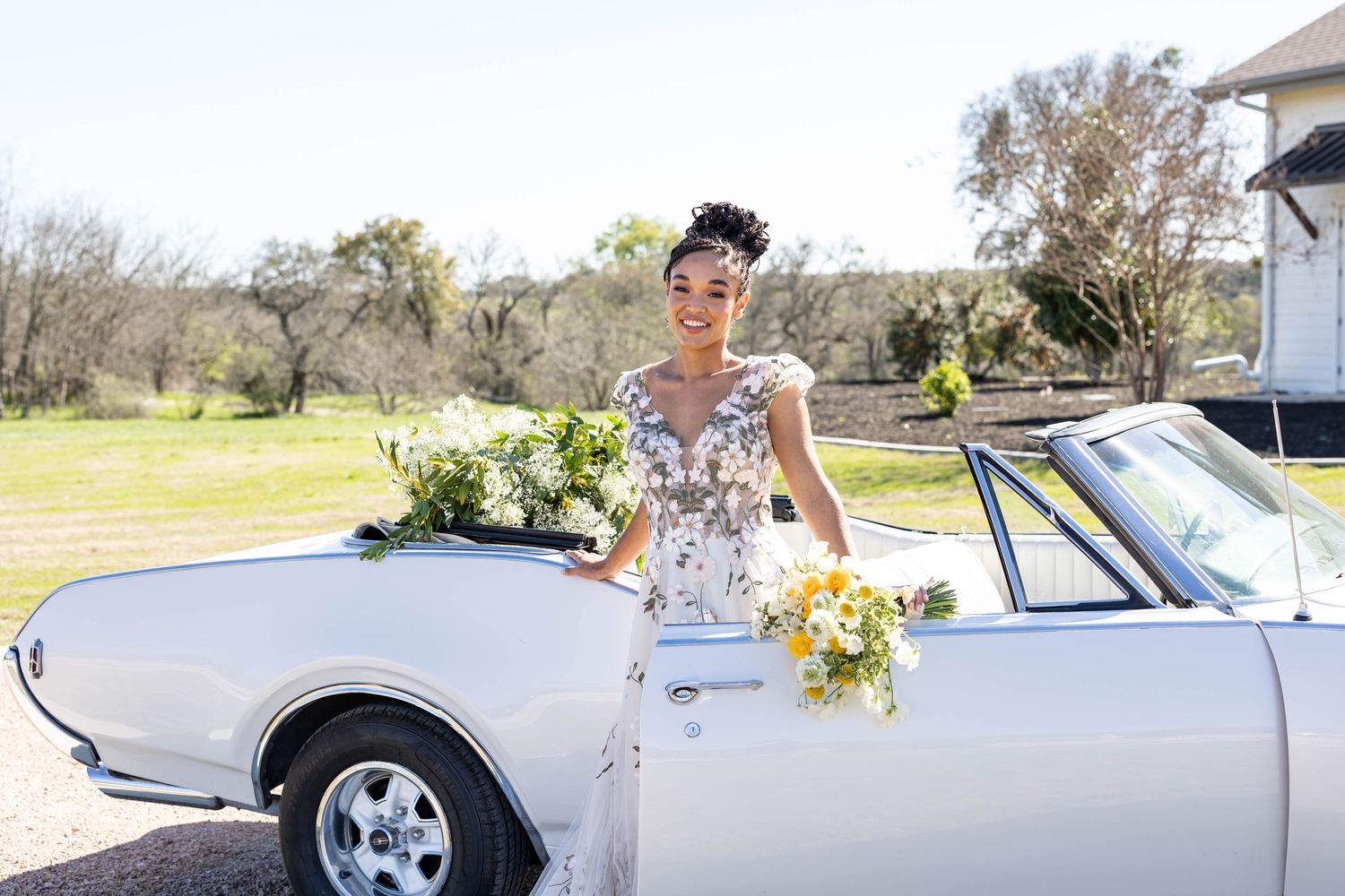 Bride with bouquet posing next to vintage white car decorated with flowers.