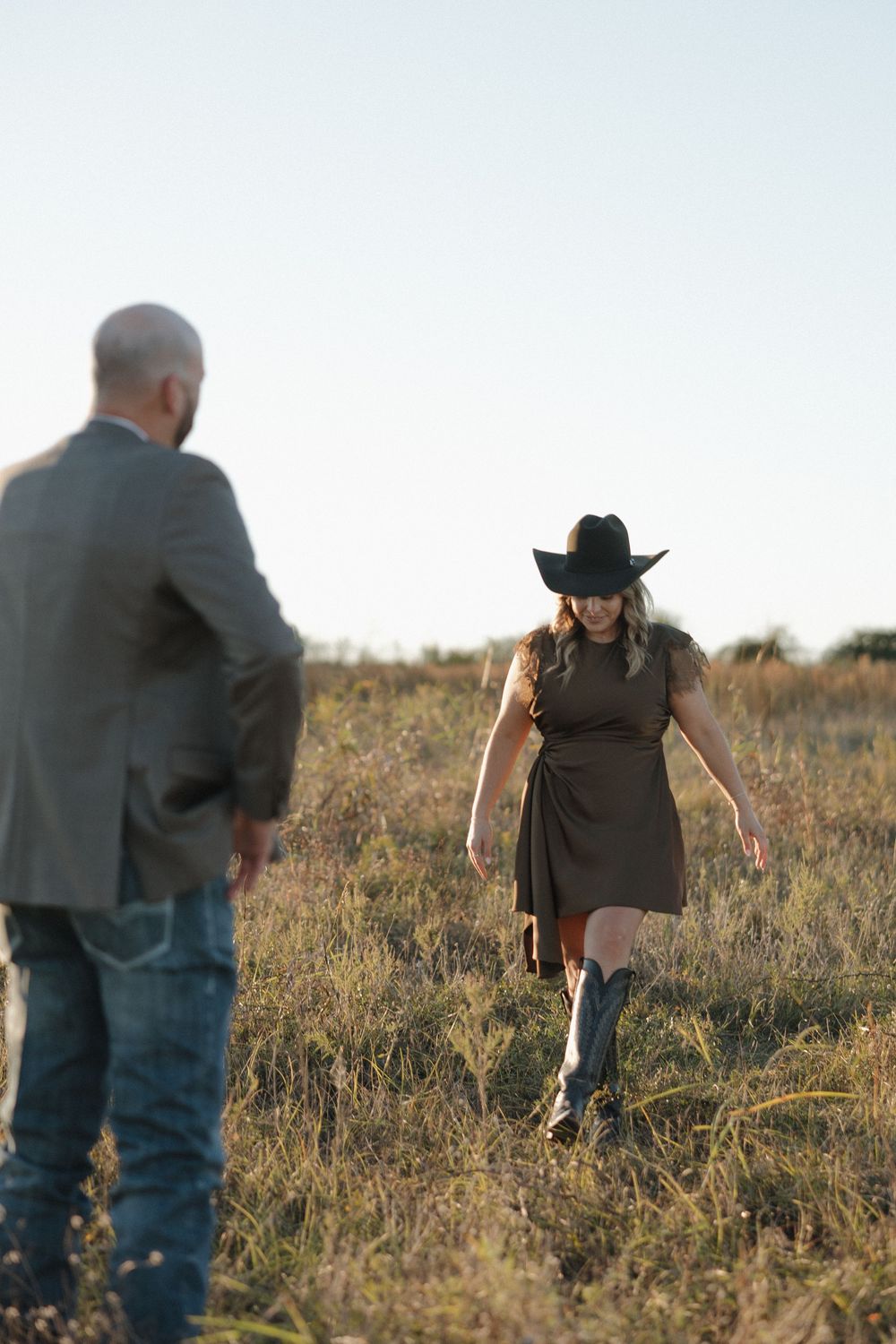 Two people in a grassy field at dusk, with one wearing a black dress, cowboy hat and boots walking through tall grass.