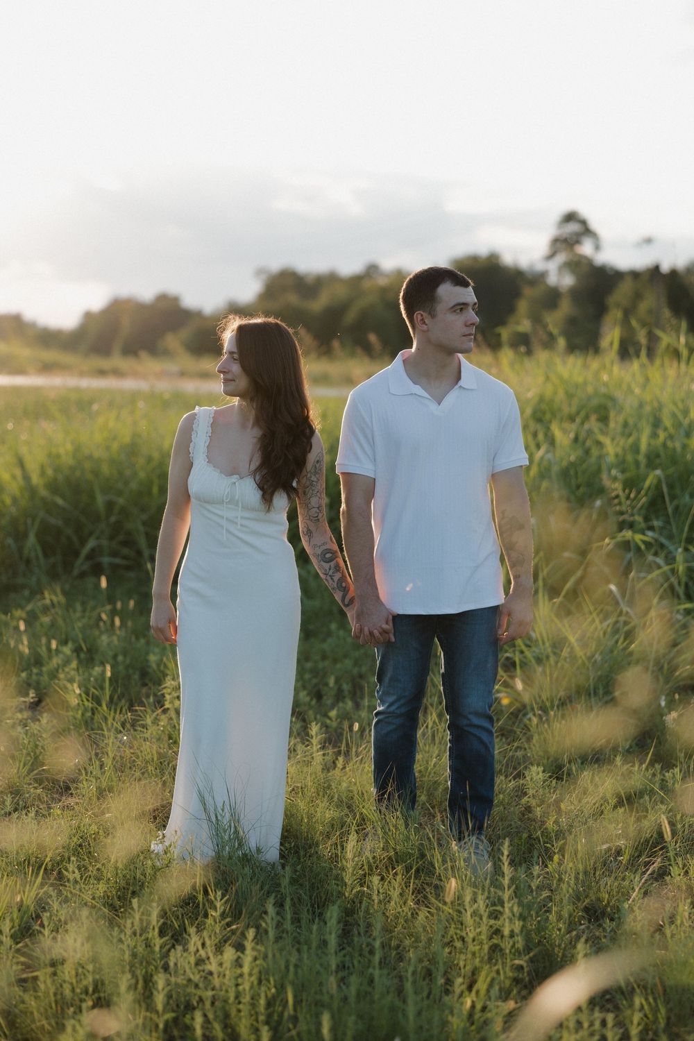 A couple holds hands while walking through a sunlit meadow at golden hour, wearing white and casual attire.
