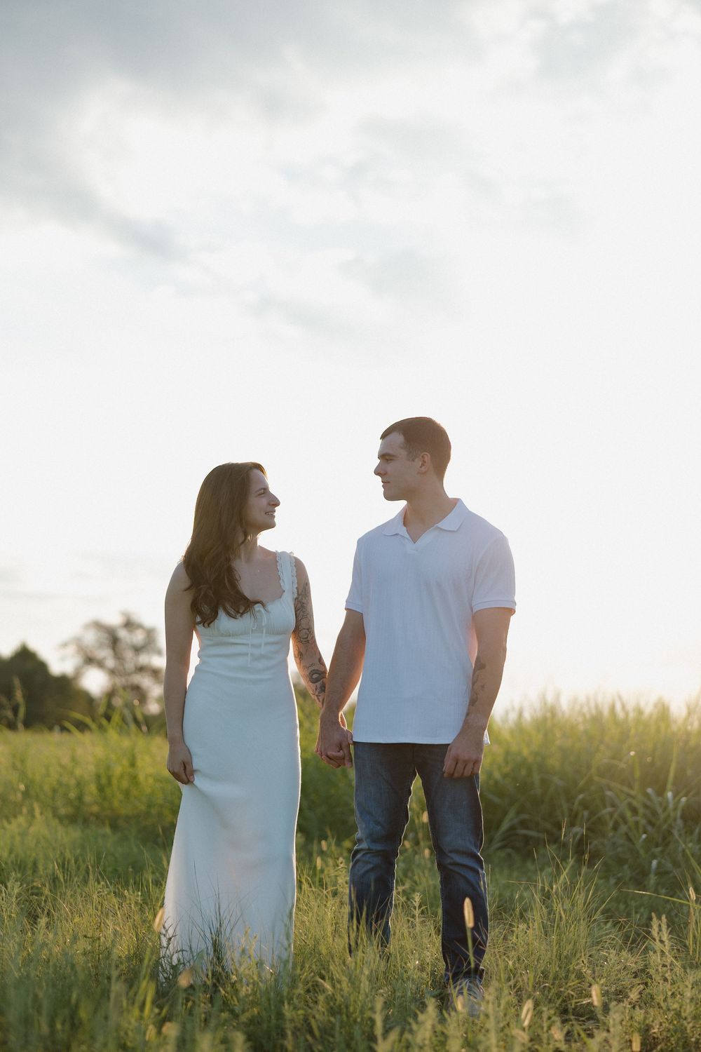 A couple walks together in a grassy meadow at sunset, wearing casual white and light blue outfits.