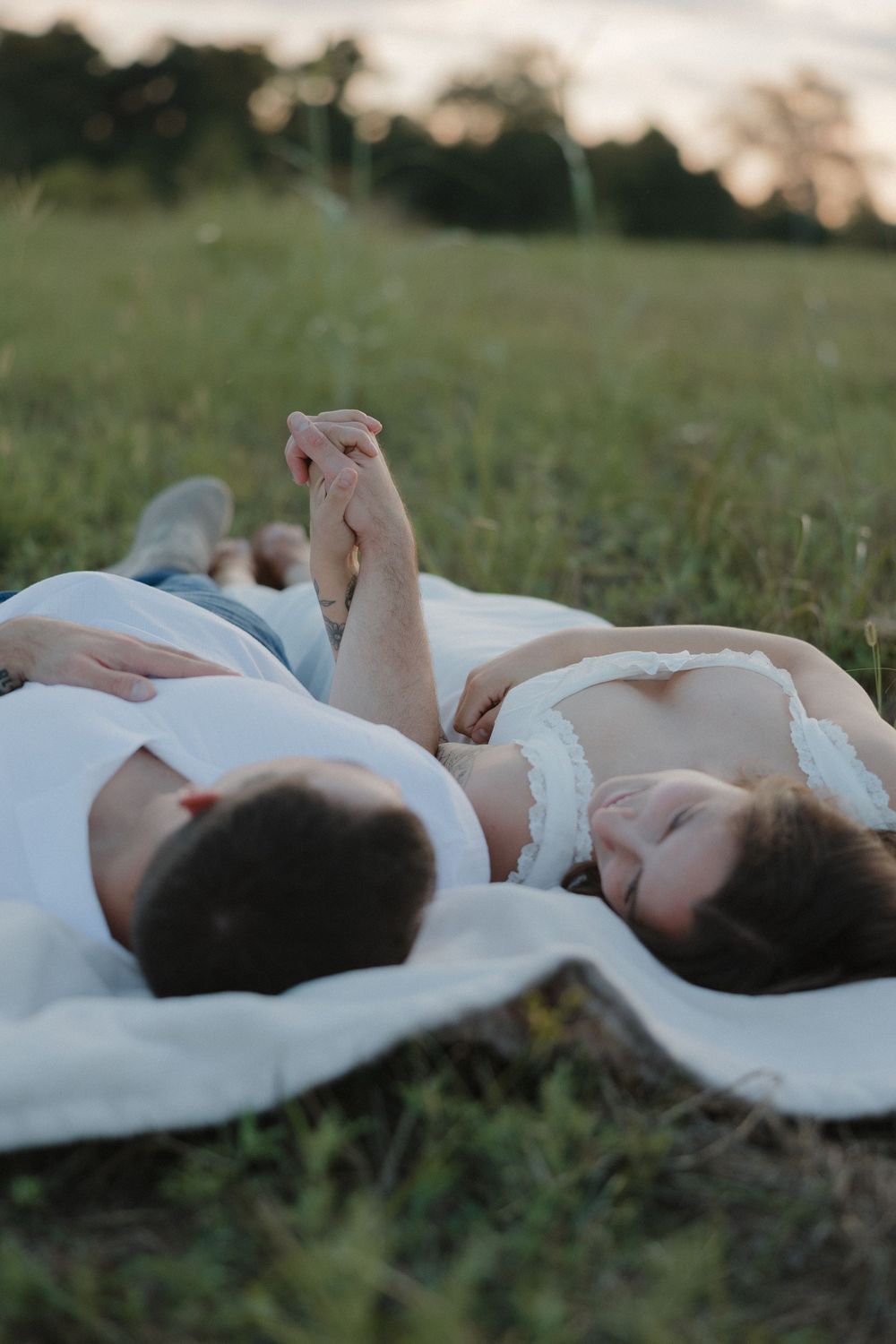Two people lying on a white blanket in a grassy field during golden hour.