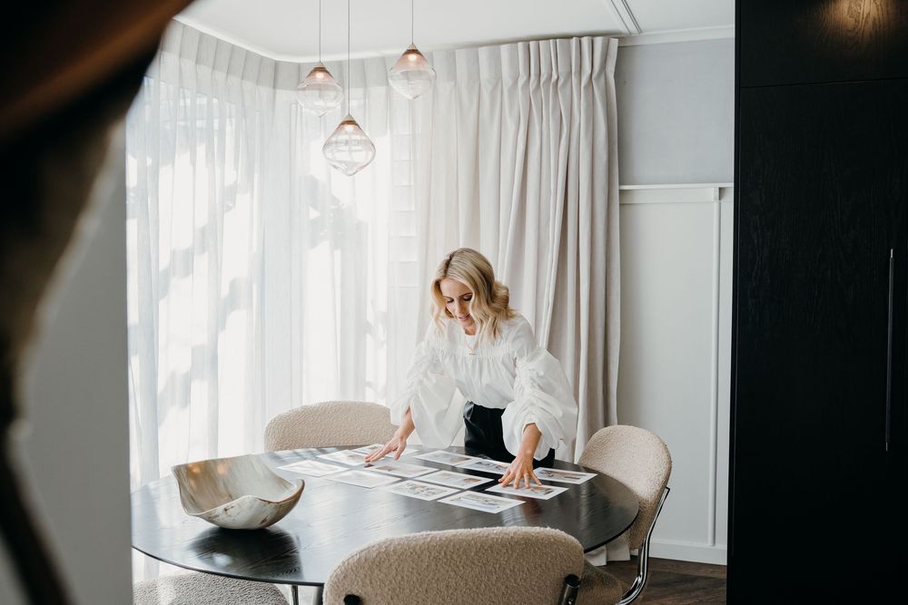 A person in white works at a modern dining table with beige chairs and pendant lights in a bright minimalist space.