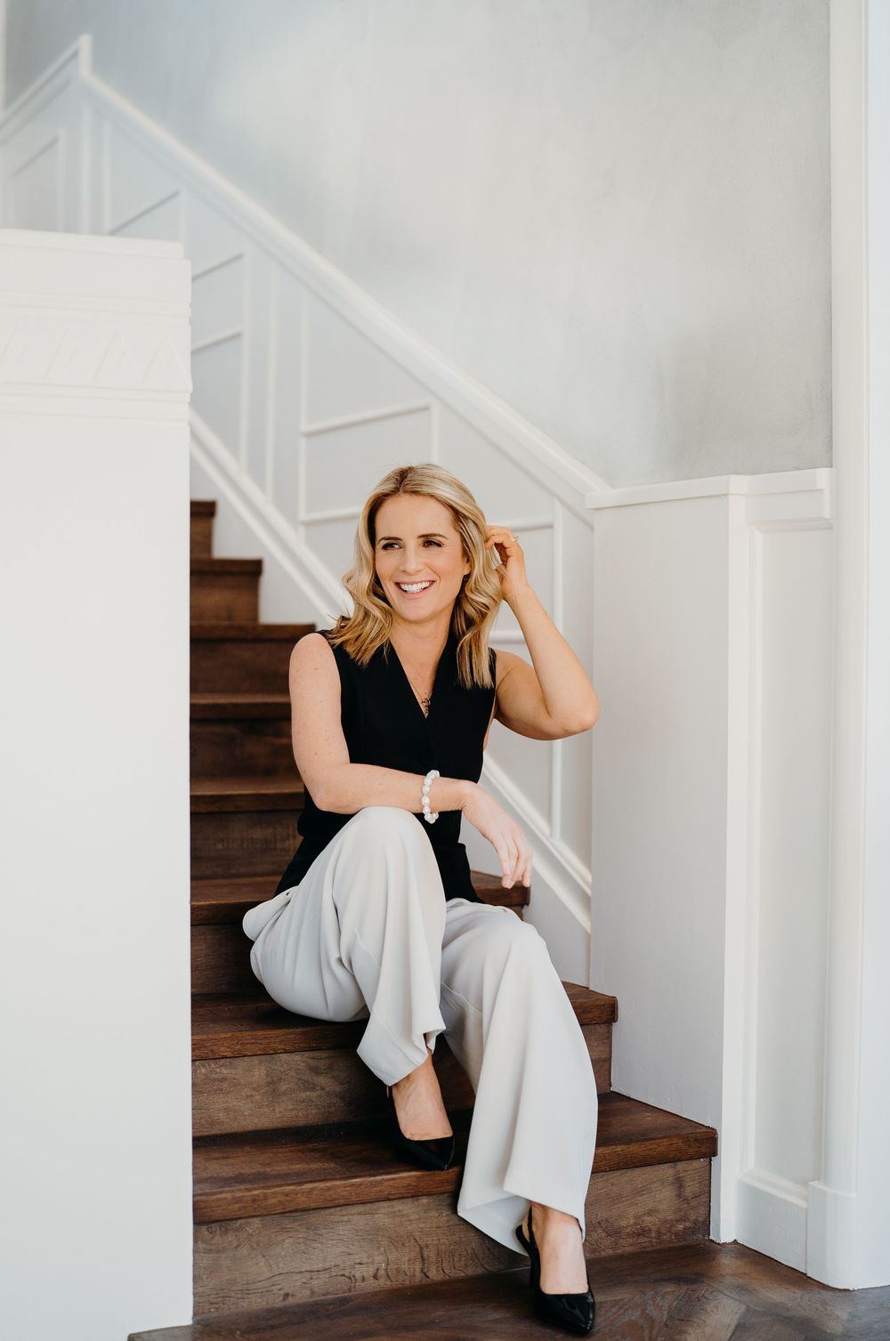 A figure in a black top and light pants sits casually on wooden stairs against a white wall interior.