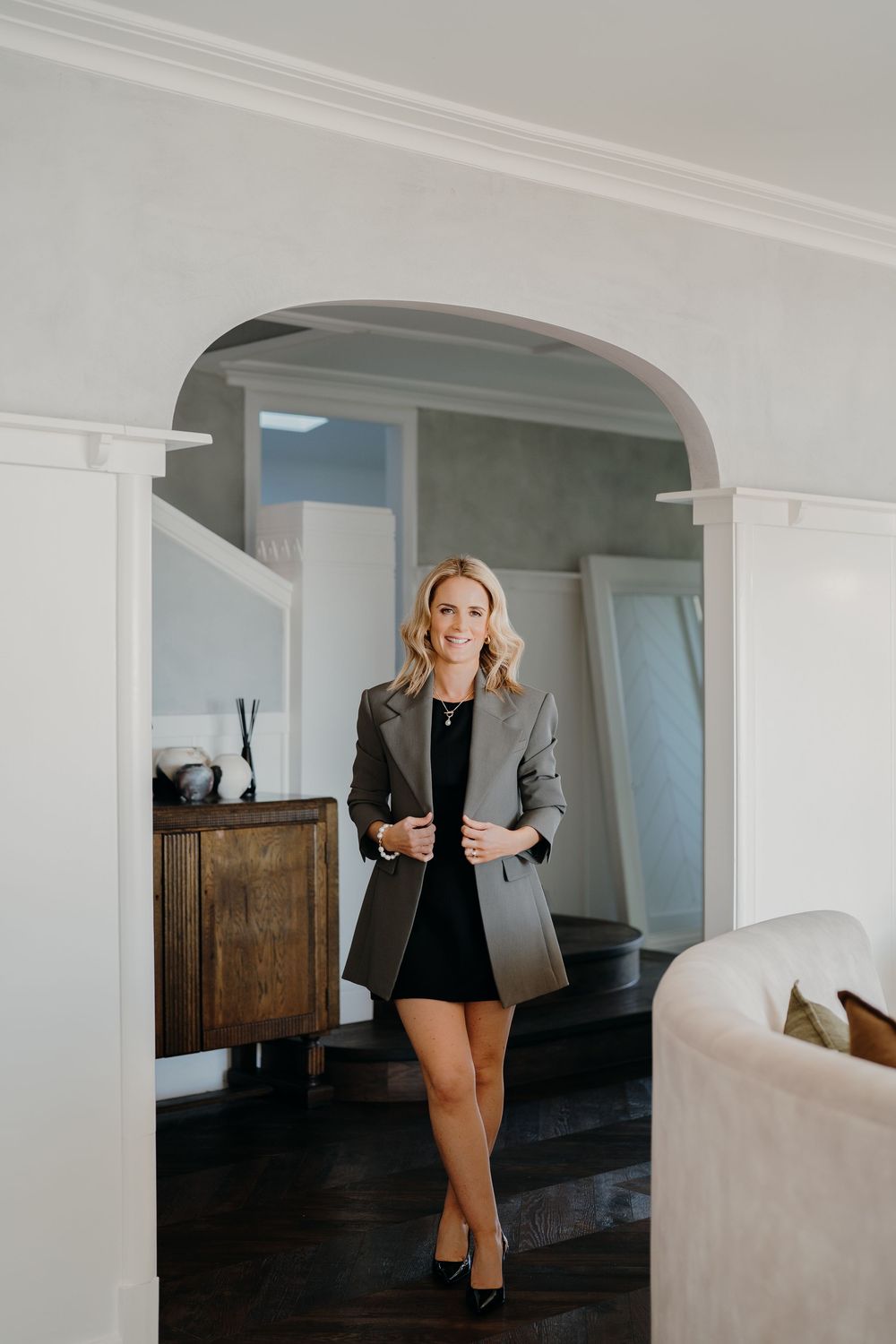 A person in a black blazer and shorts poses in a modern minimalist white room with arched doorways.