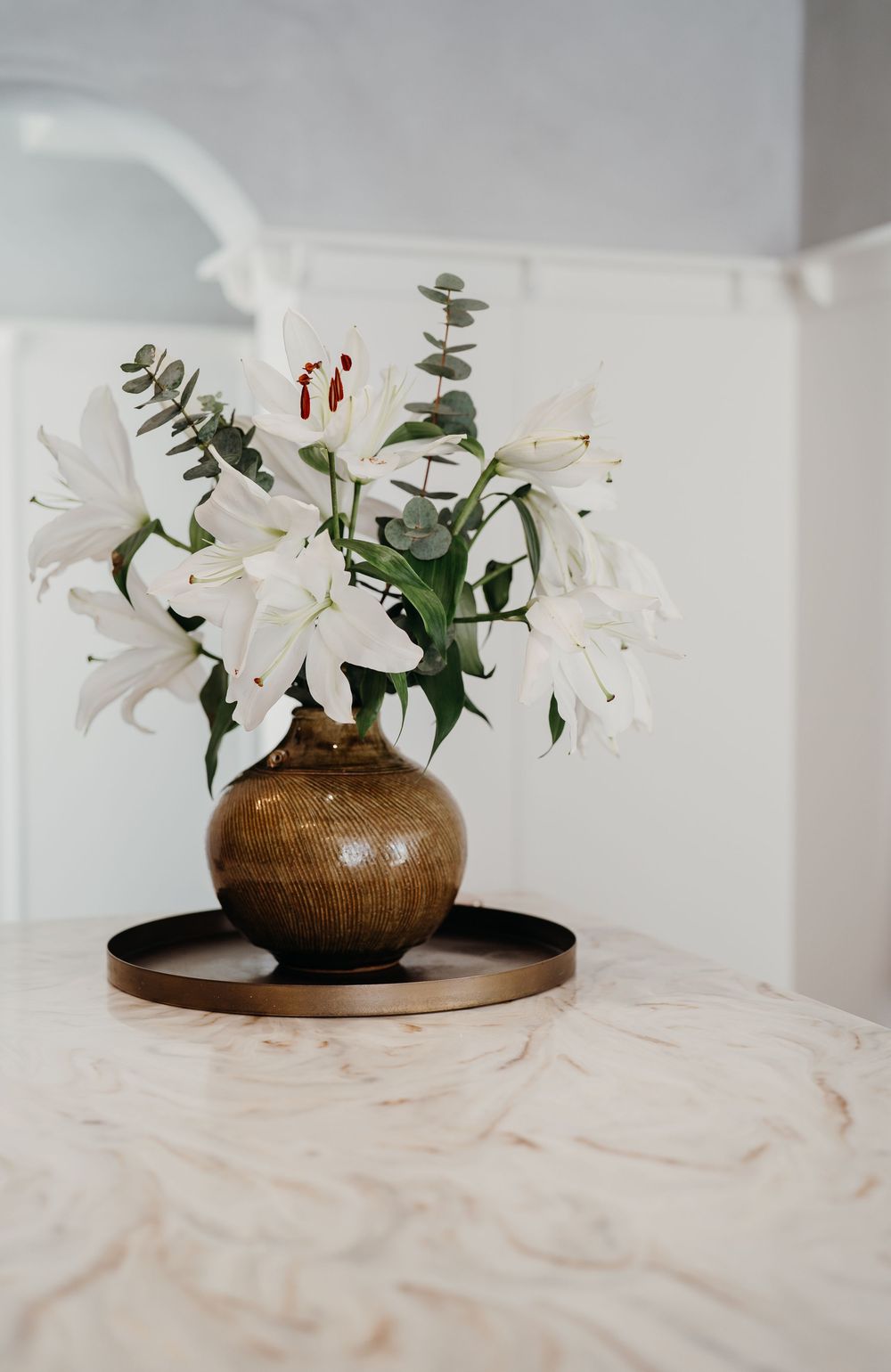White lilies and eucalyptus arranged in a brown ceramic vase on a marble surface create an elegant centerpiece display.