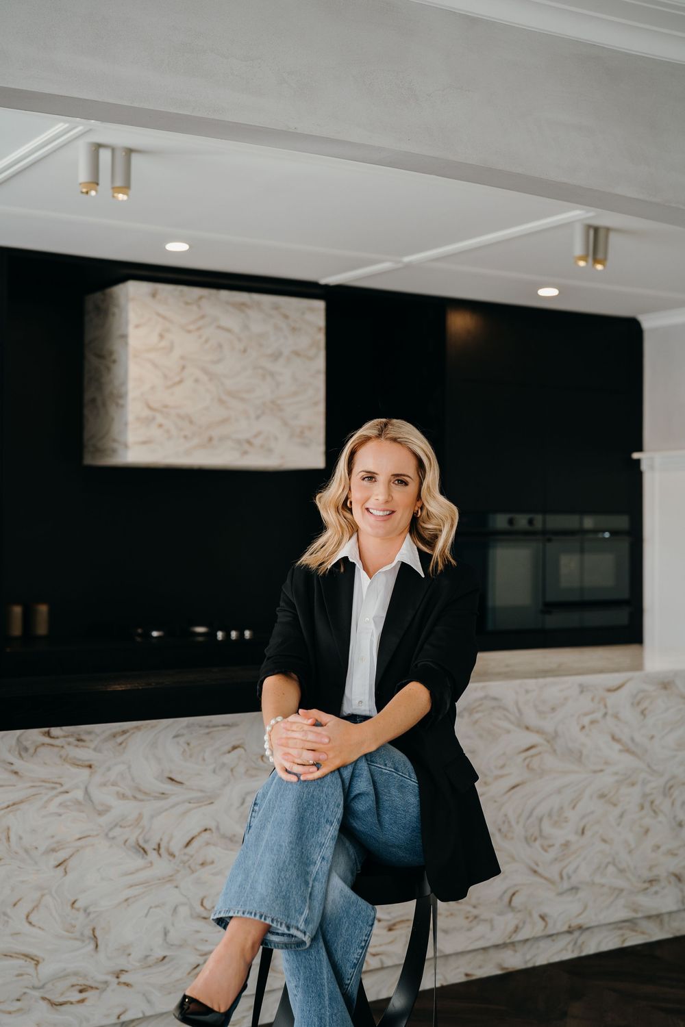Professional wearing black blazer and jeans sitting casually in modern office space with marble wall details.