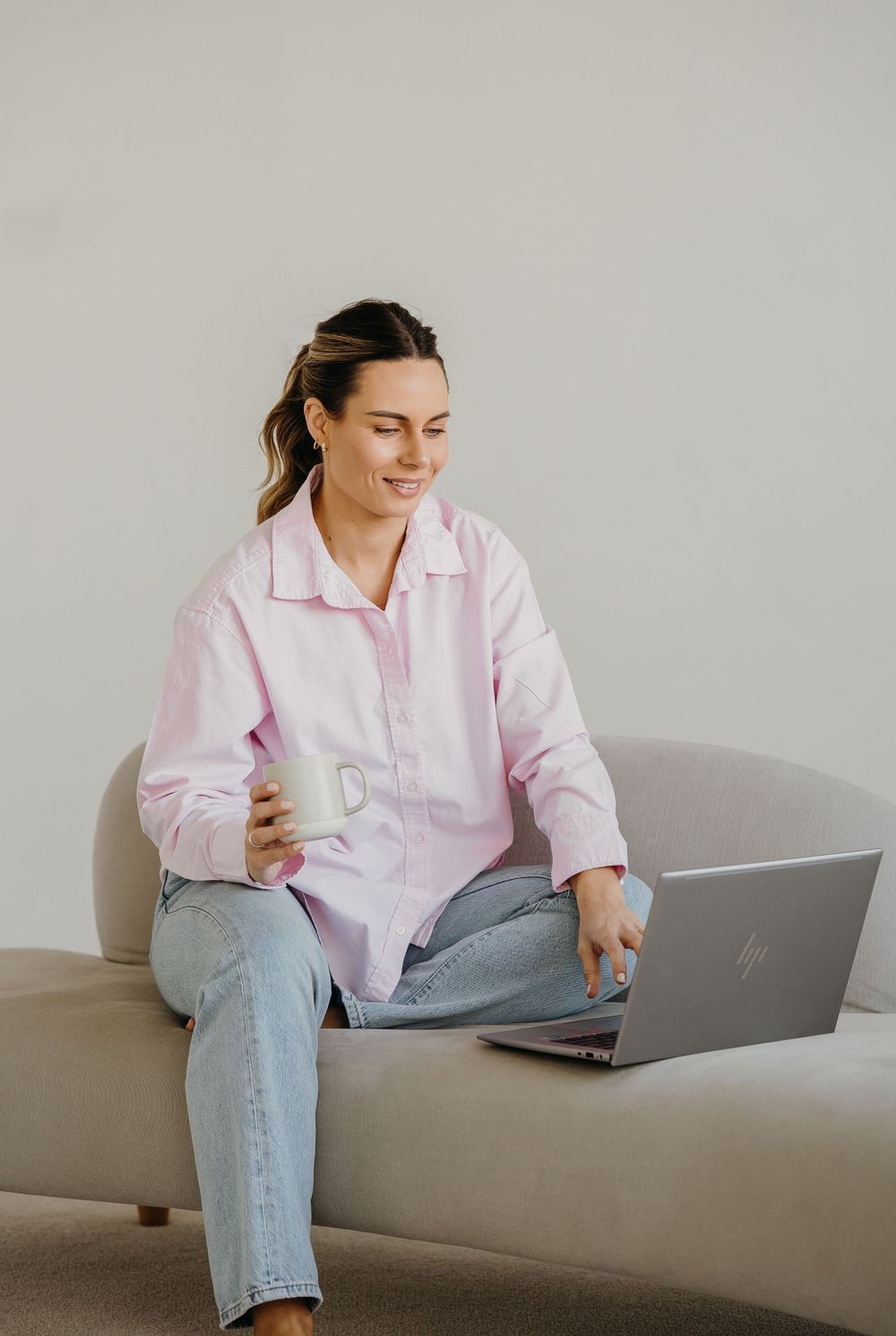 Person in pink button-down shirt and jeans works on laptop while relaxing on gray couch.