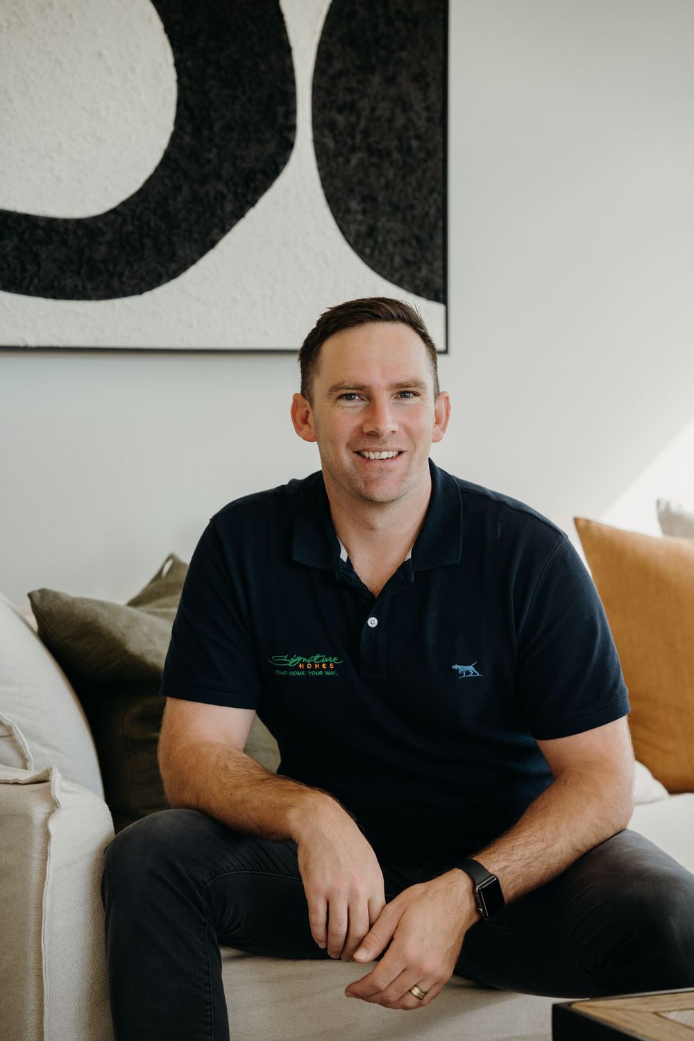 Someone sits casually on a light-colored couch wearing a navy polo shirt against a white wall with abstract art.