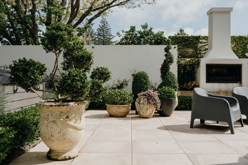 Modern outdoor patio with white walls, potted topiary plants, and dark lounge chair next to an outdoor fireplace.