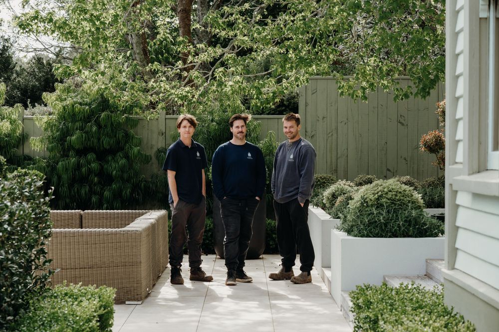 A group of people in dark clothing stand outside on a porch near greenery and white columns.
