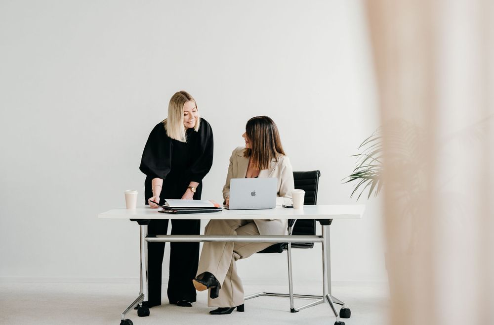 Two professionals collaborating at a modern white desk with a laptop and office supplies in a minimalist workspace.