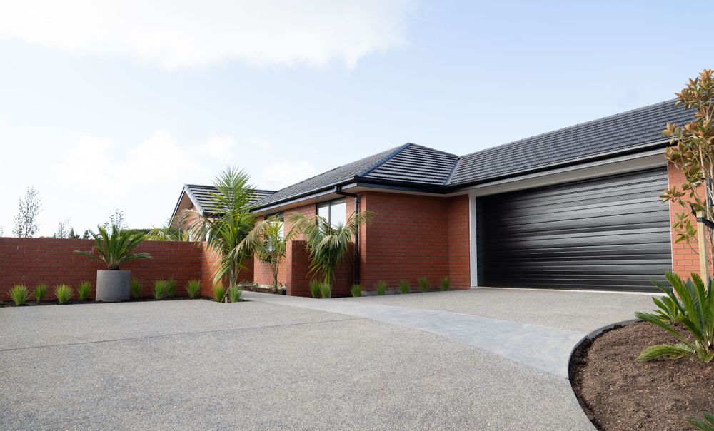 Modern brick home with black garage door and tropical landscaping features a circular driveway under cloudy skies.