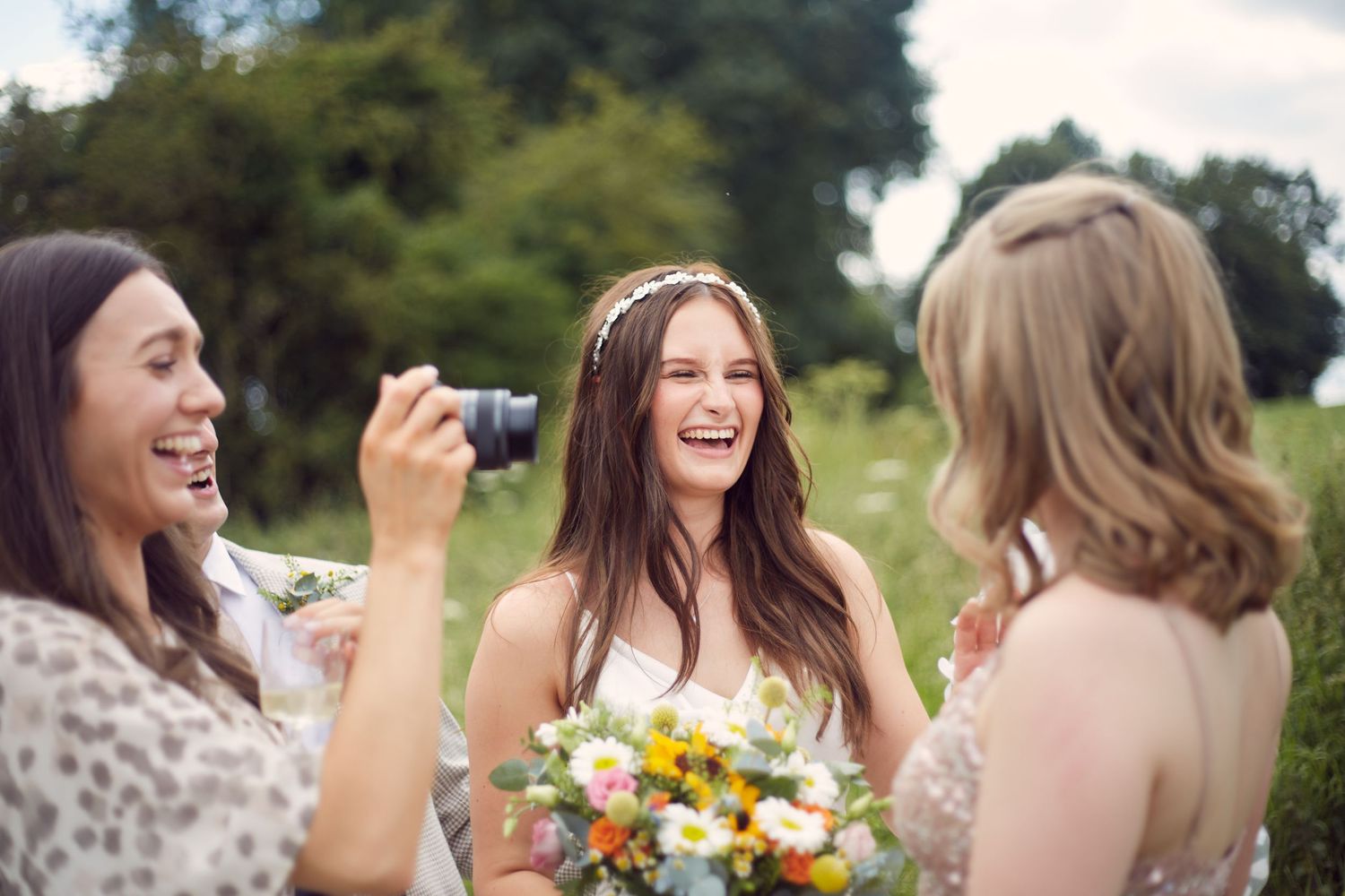 Friends laugh together while holding flowers and a camera at an outdoor wedding celebration.