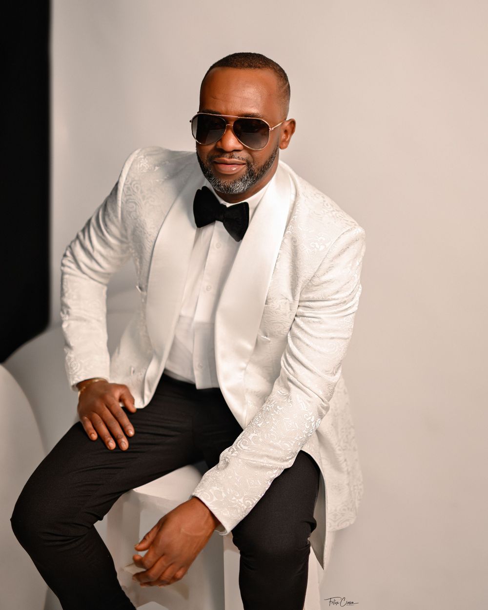 A stylishly dressed person in a white dinner jacket, black bow tie, dark pants and sunglasses poses for a formal portrait.