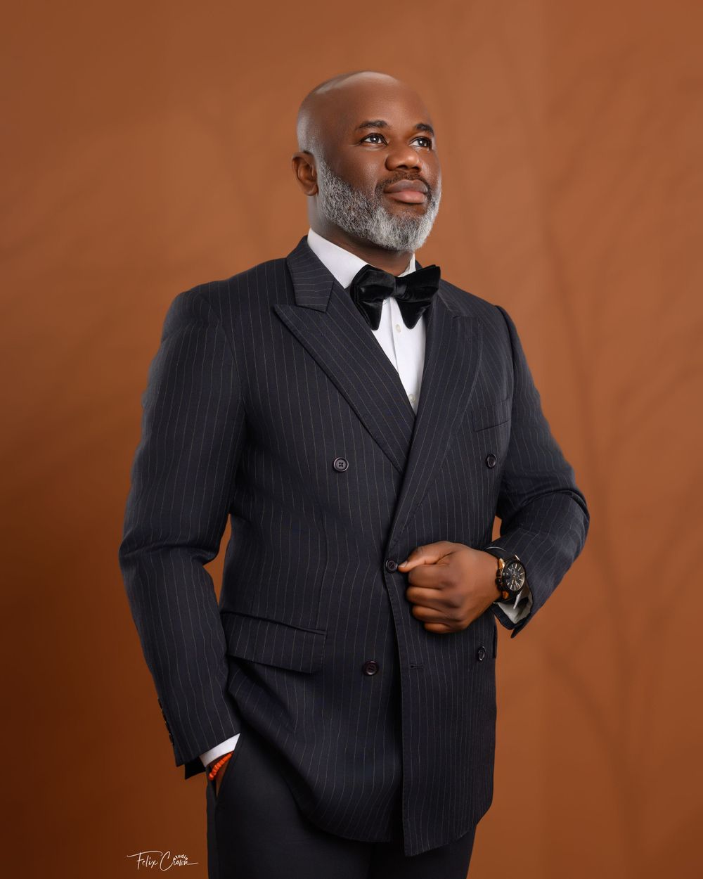 An elegant man in a pinstriped double-breasted suit and bowtie smiles while posing against a brown backdrop.