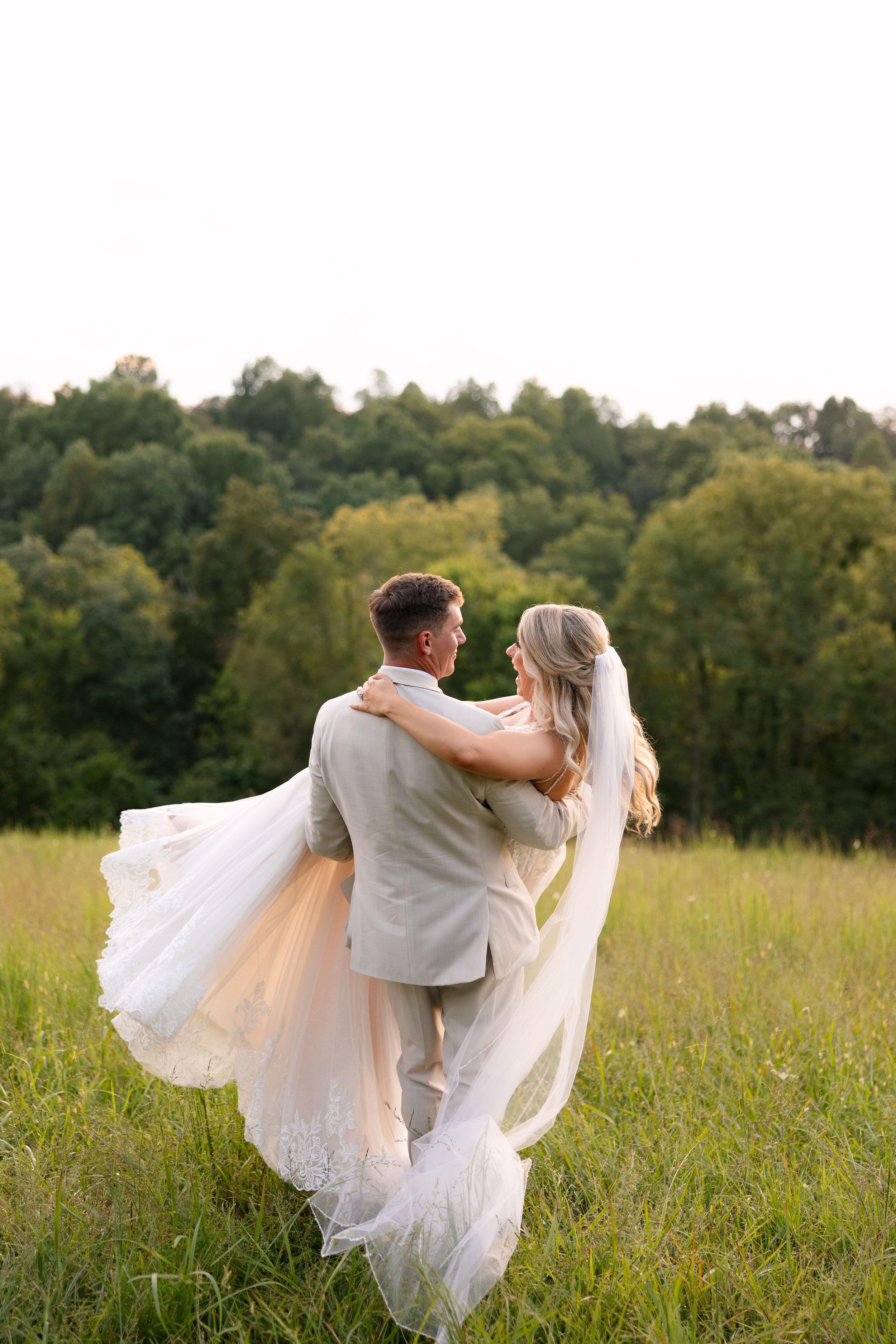Groom lifting bride in white gown and veil in a sunlit green meadow with trees in the background.