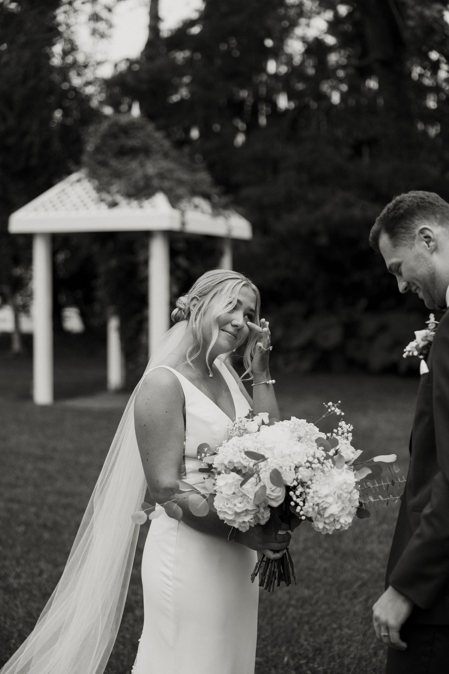 Emotional bride in white gown holding floral bouquet wipes tear as groom smiles beside her at outdoor wedding ceremony.