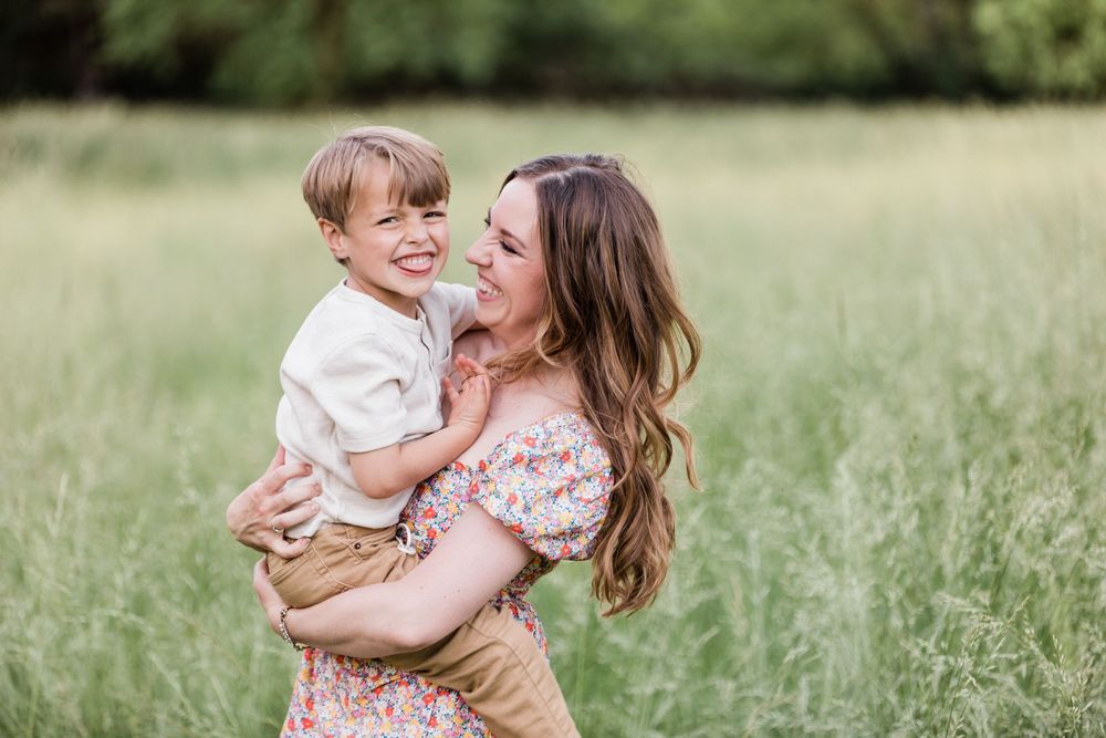 Mother and child share affectionate moments in sunlit field while wearing floral and neutral colored outfits.