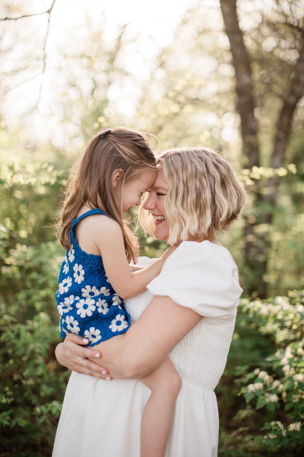 A tender moment captured in sunlit woods as parent and child share an intimate embrace in white and blue floral dresses.