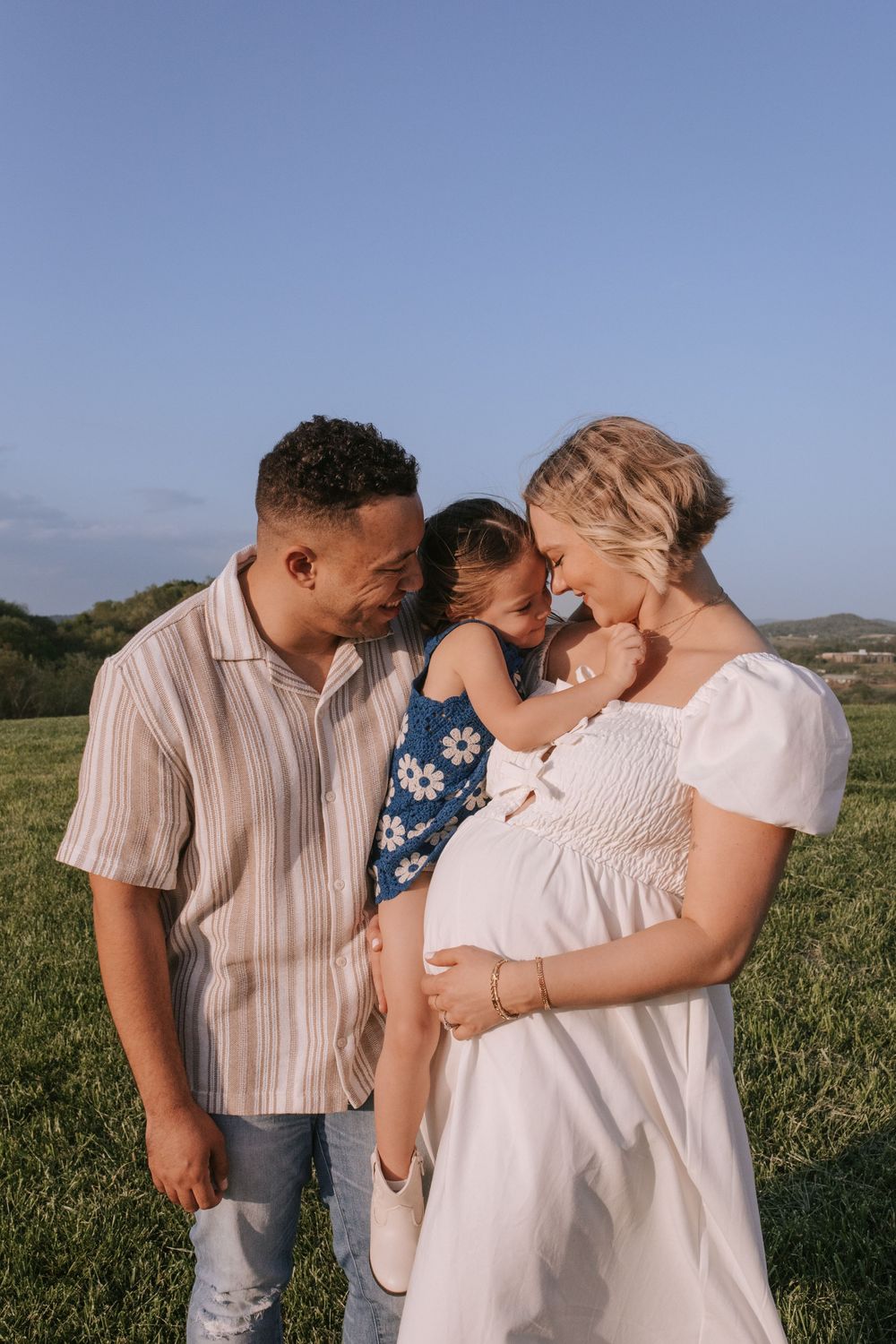 Family enjoying tender outdoor moment together in grassy field under blue sky.