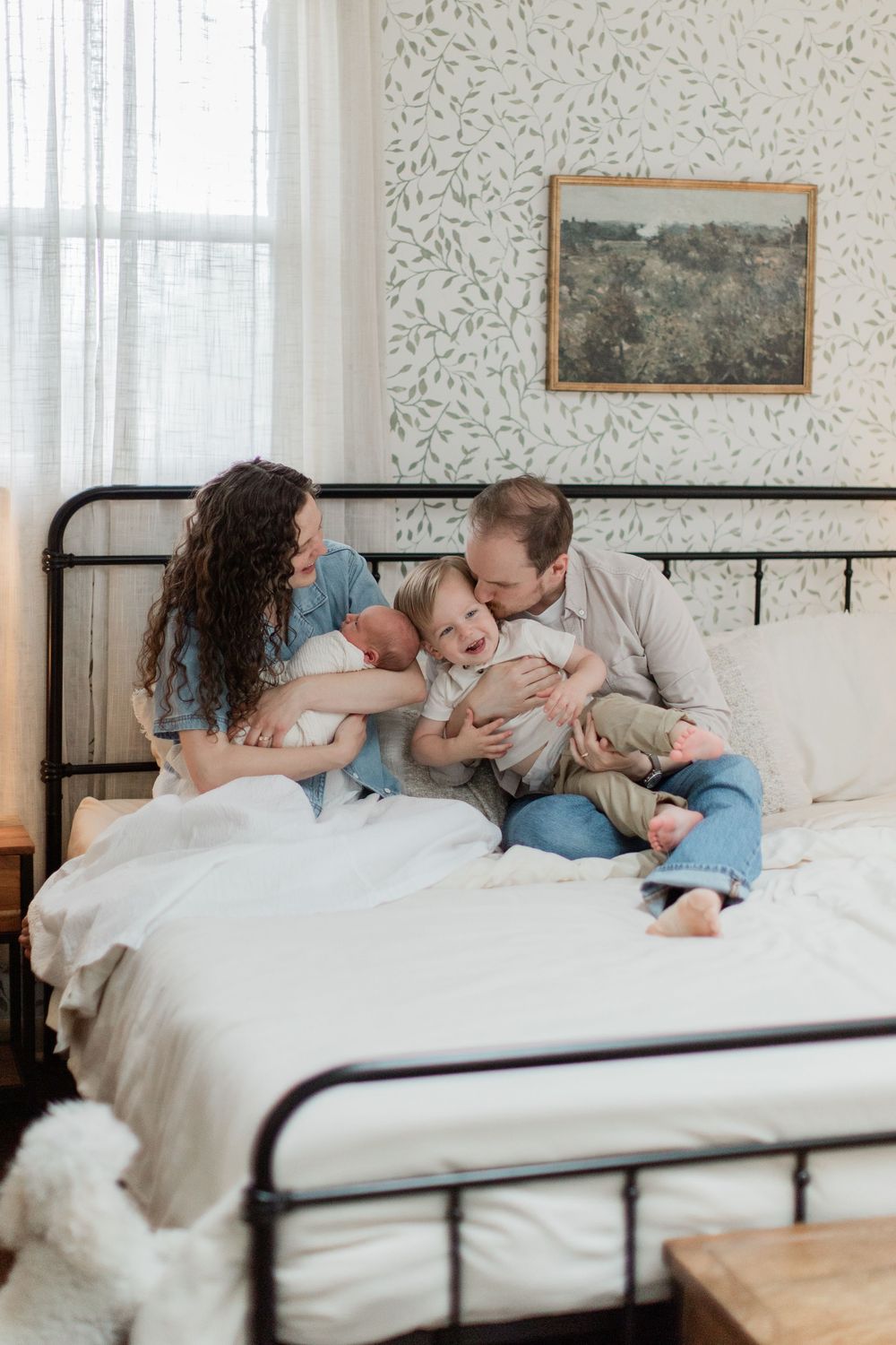 A family snuggles together warmly on a white bed with black iron frame in a light-filled bedroom.