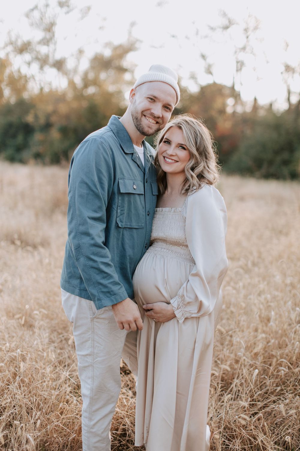 A couple stands together in a golden field during sunset, with her wearing a flowing dress highlighting her pregnancy.