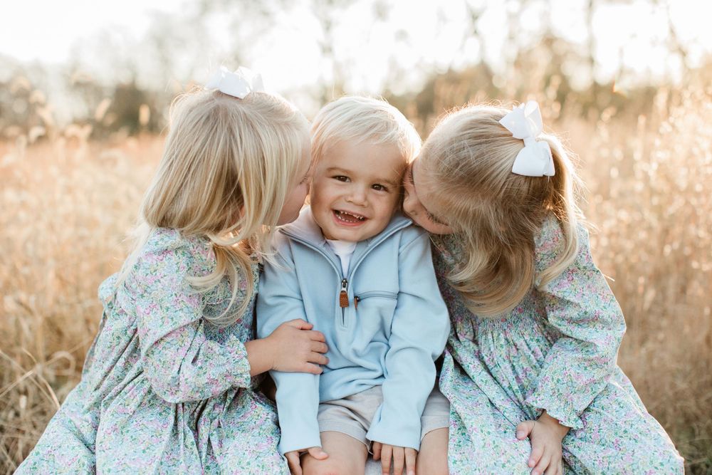 Two children in matching light blue and floral dresses laugh and embrace while playing outdoors in a golden field.