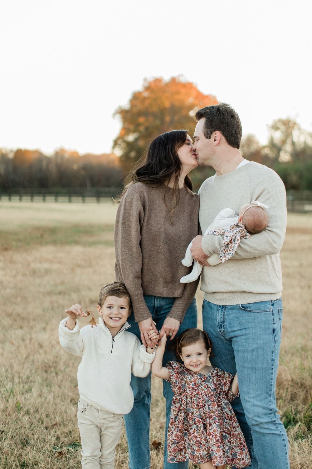 A family of five stands close together in a fall field at sunset wearing cozy sweaters and casual attire.