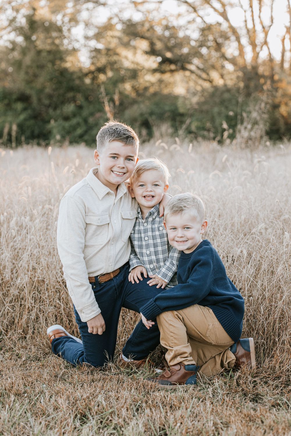 Three siblings in coordinating outfits pose together in a field of tall beige grass during golden hour.