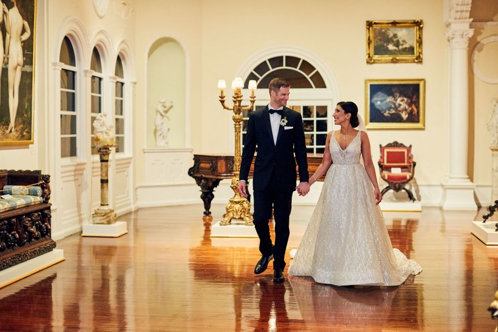 Couple walks through elegant ballroom with chandelier and arched windows.