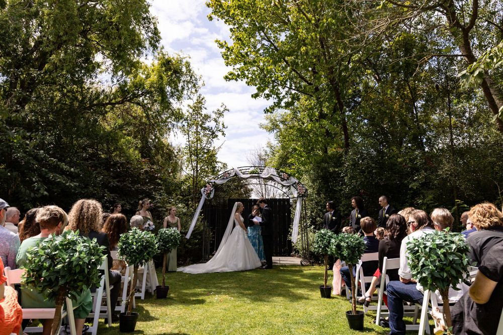 An outdoor wedding ceremony in progress with guests seated on white chairs along a tree-lined aisle with floral decorations.