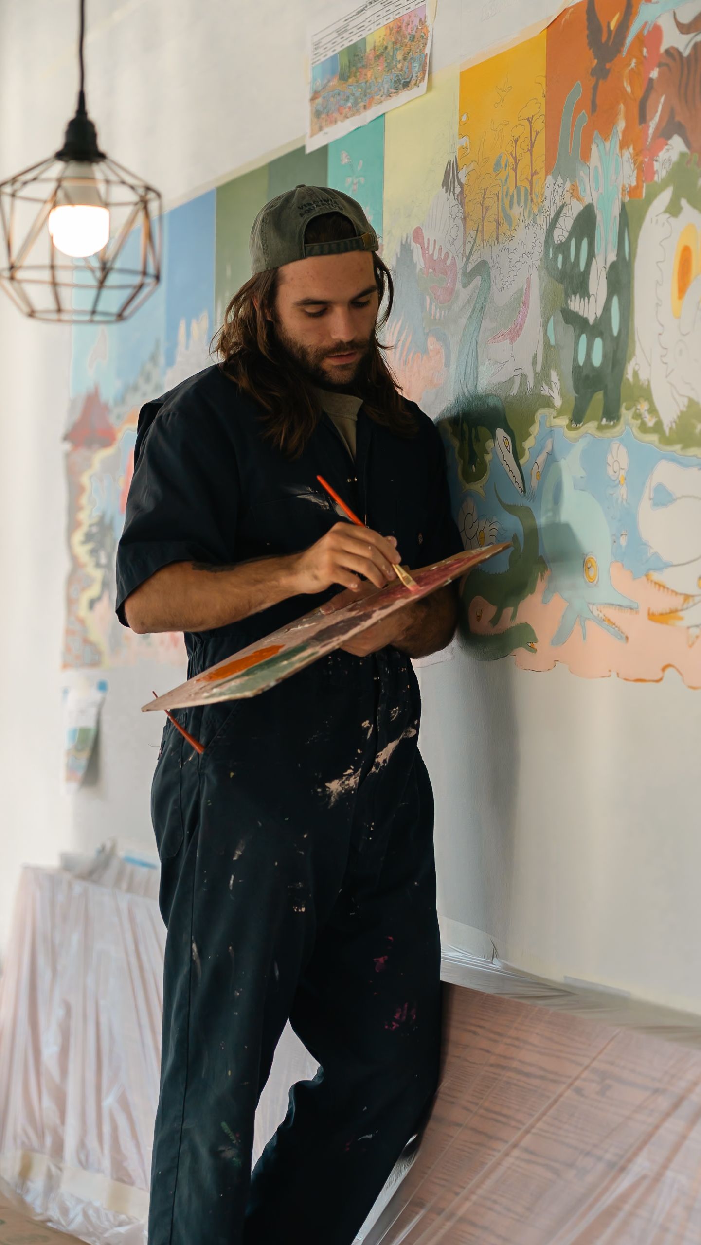 Artist in dark clothing sketches in a notebook against a colorful wall mural background.