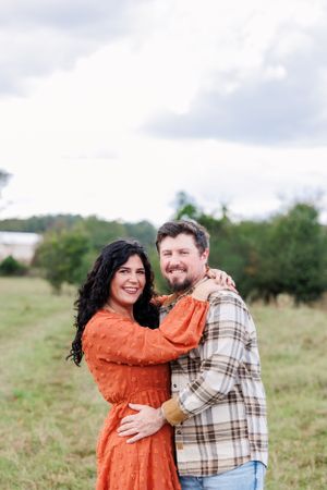 A couple embraces in an open field wearing casual fall attire on a cloudy day.