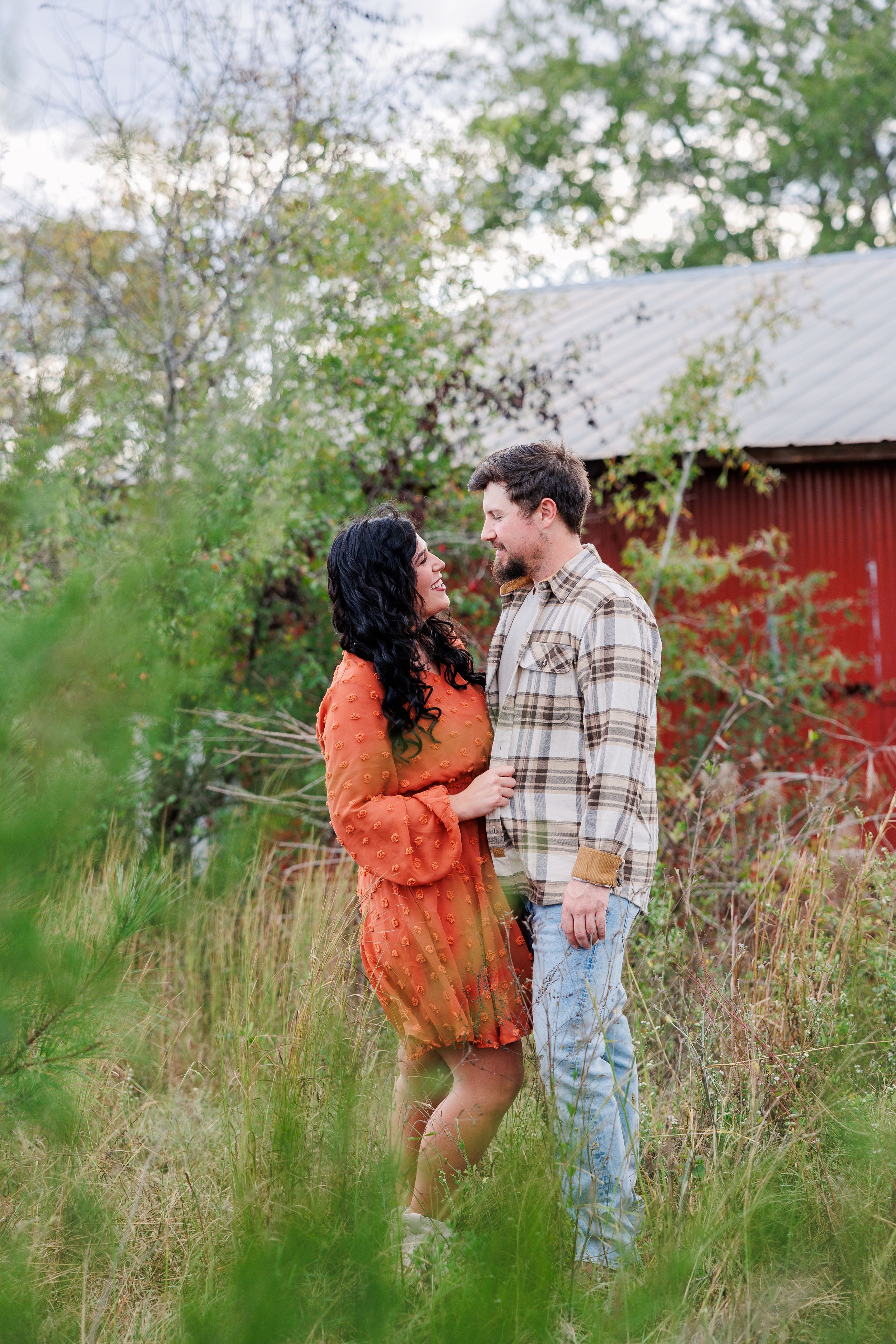 A couple poses romantically near a red barn surrounded by tall grass and autumn foliage.